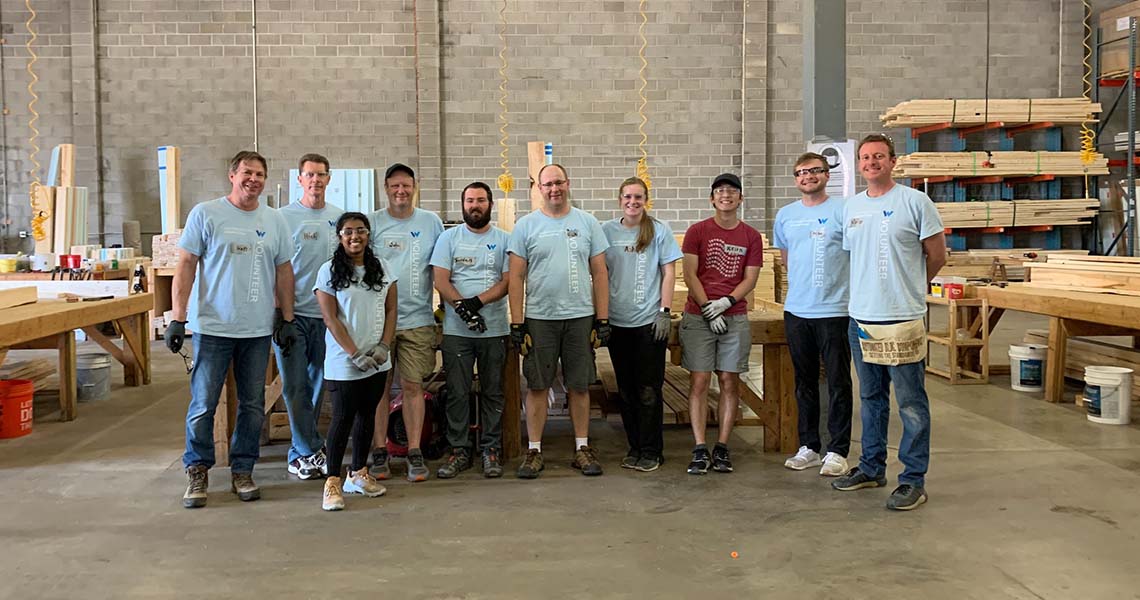 Western National employee volunteers wearing work gloves, safety goggles, and matching Western National branded t-shirts and posing for a group photo in a warehouse surrounded by lumber.