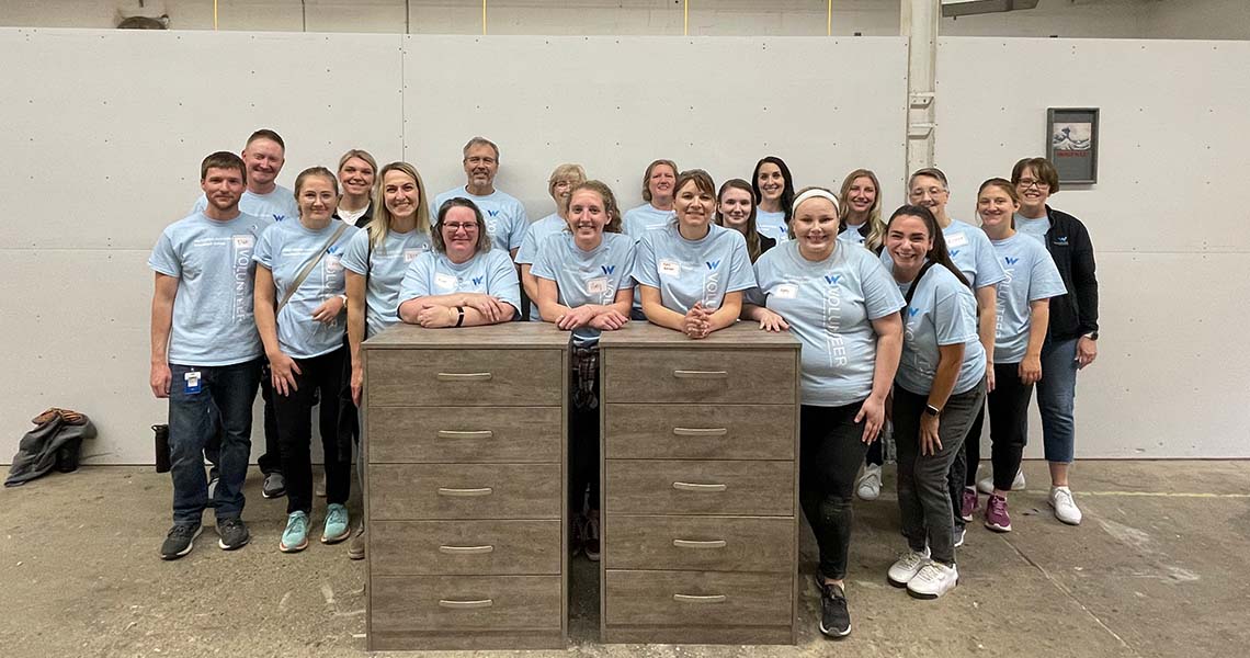 Western National employee volunteers wearing matching Western National branded t-shirts and posing for a group photo in a warehouse with wooden dressers they built during a volunteer event.